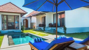 A poolside area with lounge chairs and a large umbrella, a house with a tiled roof, and a well-maintained lawn.