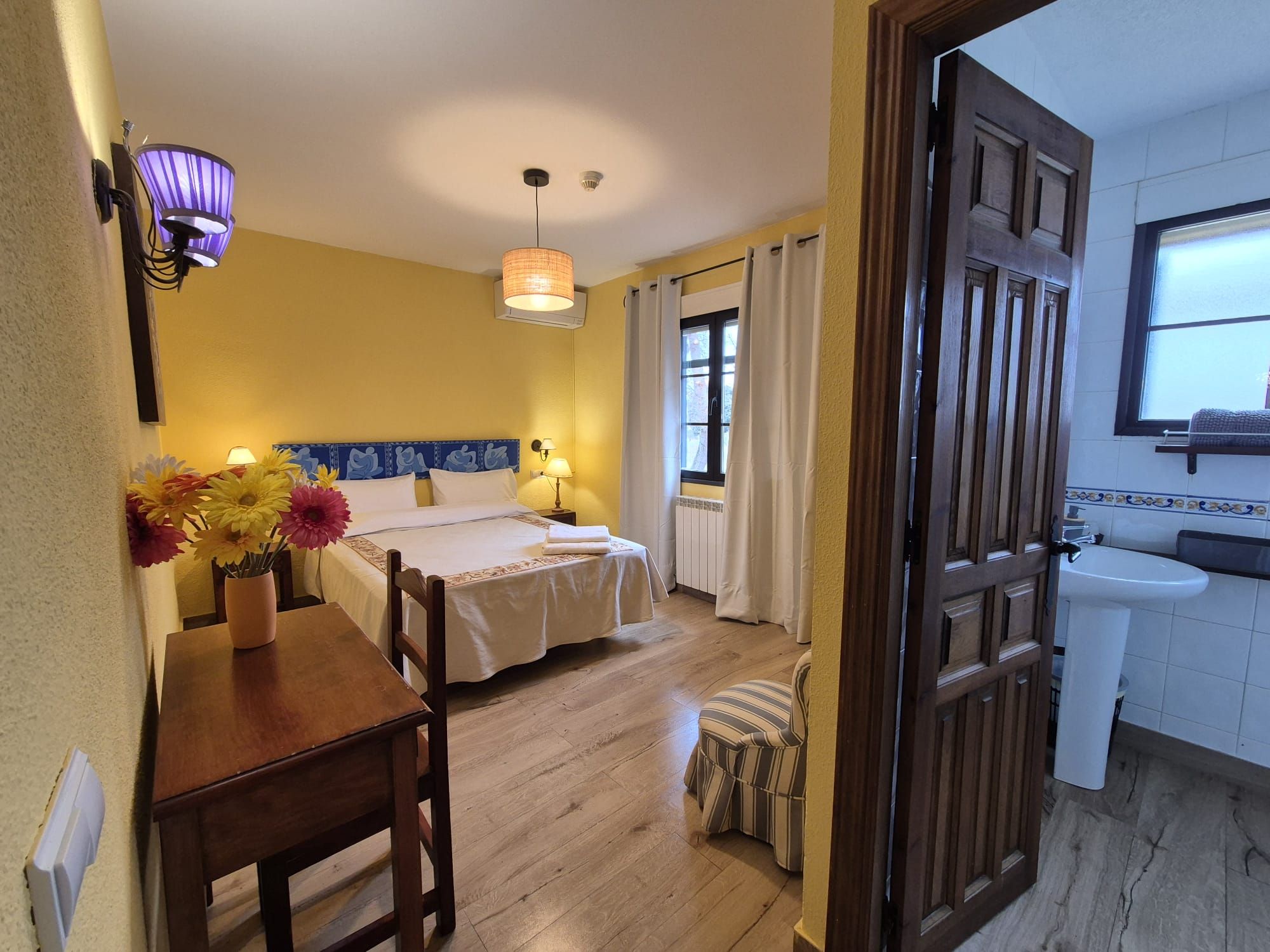 A hotel room with a bed, bedside table, flowers, a small stool, a window, and a bathroom with a sink.