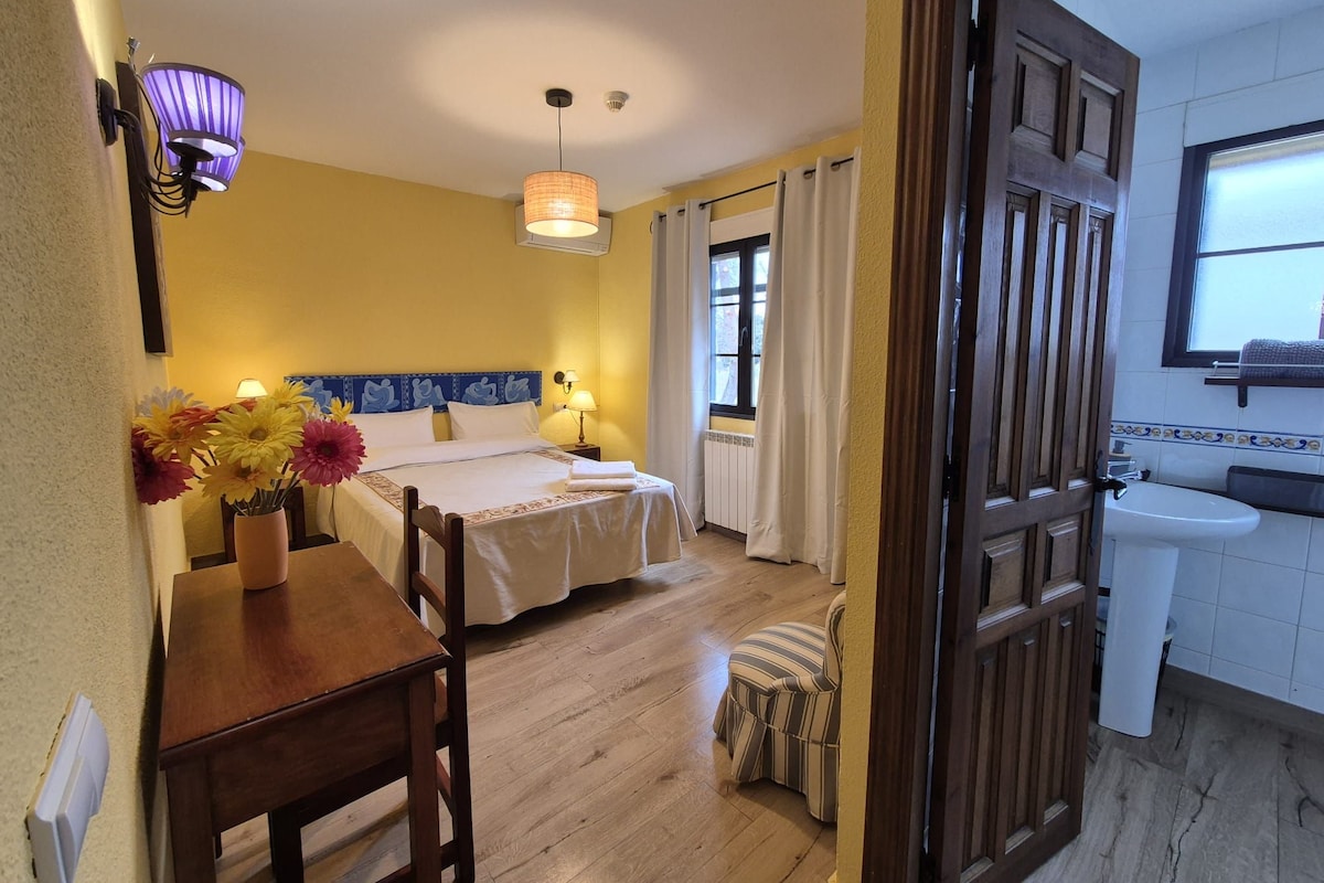 A hotel room with a bed, bedside table, flowers, a small stool, a window, and a bathroom with a sink.