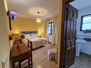 A hotel room with a bed, bedside table, flowers, a small stool, a window, and a bathroom with a sink. - La Casona de Tamaya (Tamajón)