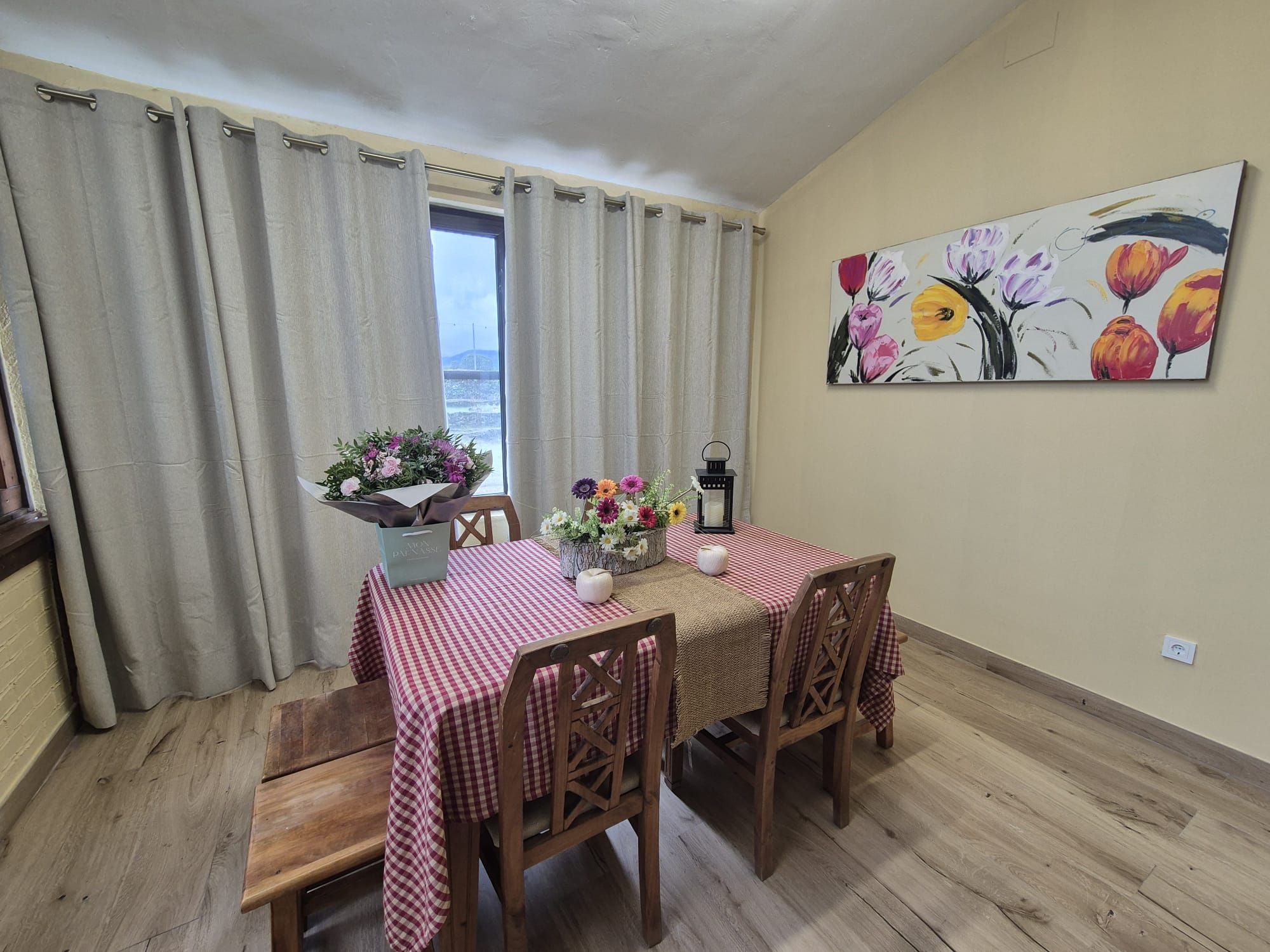 A dining area with a checkered tablecloth, wooden chairs, a vase with flowers, and a painting on the wall.