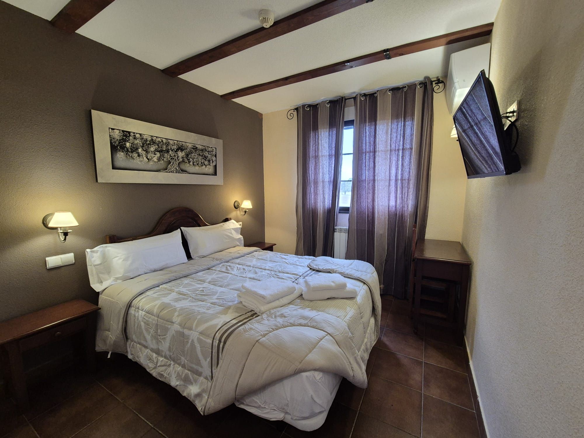 Una habitación de hotel con cama, lámparas de noche, televisor montado y ventana con cortinas.