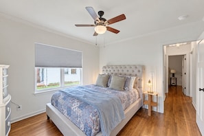 A bedroom with a bed, a ceiling fan, a window with blinds, and a hallway leading to another room.