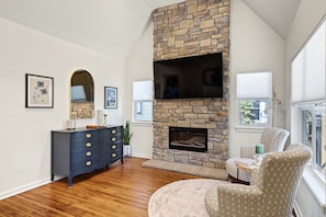 A living room with a stone fireplace, a flat-screen TV, a dark blue dresser, and patterned armchairs.