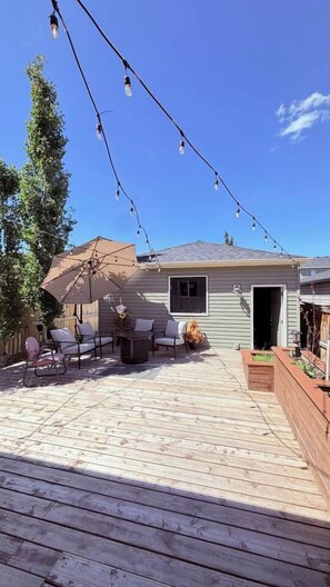 Una terraza de madera con mobiliario exterior, luces colgantes y una casa al fondo.