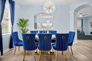 Un comedor con una mesa redonda, sillas tapizadas en azul, una araña y una planta en maceta.