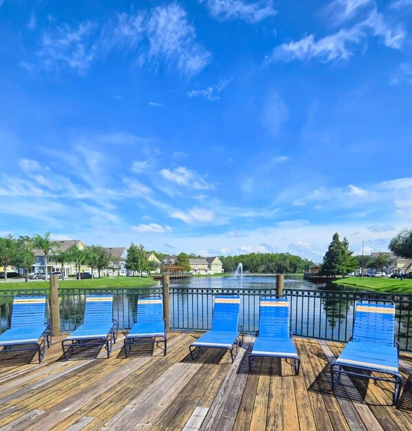Une terrasse en bois avec des chaises longues bleues donnant sur un lac paisible, le tout entouré d’immeubles d’habitation.