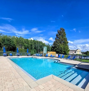 Une piscine entourée d’une clôture et d’arbres, avec des chaises et des parasols bleus.