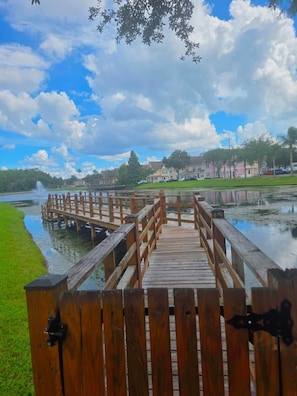Une promenade en bois qui mène à un plan d’eau agrémenté d’une fontaine, entouré d’immeubles résidentiels et de verdure.