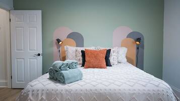 Un lit recouvert d’une courtepointe blanche et agrémenté d’oreillers décoratifs, une porte blanche et un mur orné de motifs abstraits.