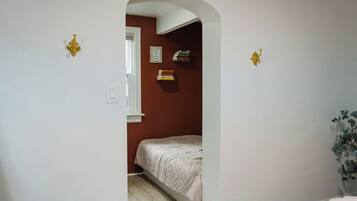 A narrow hallway with a bed in a room at the end, a white door, and a small plant on the right.