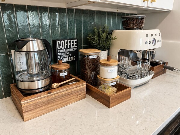 Een koffiehoek met een koffiezetapparaat, potten koffiebonen, suiker, honing en een bordje met de tekst 'COFFEE A WARM AND DELICIOUS ALTERNATIVE TO EVERYBODY'S PROBLEMS'.
