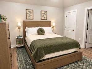 Un dormitorio ordenado con cama de madera, dos lámparas de pared, una mesita de noche con una planta y una puerta a la derecha.