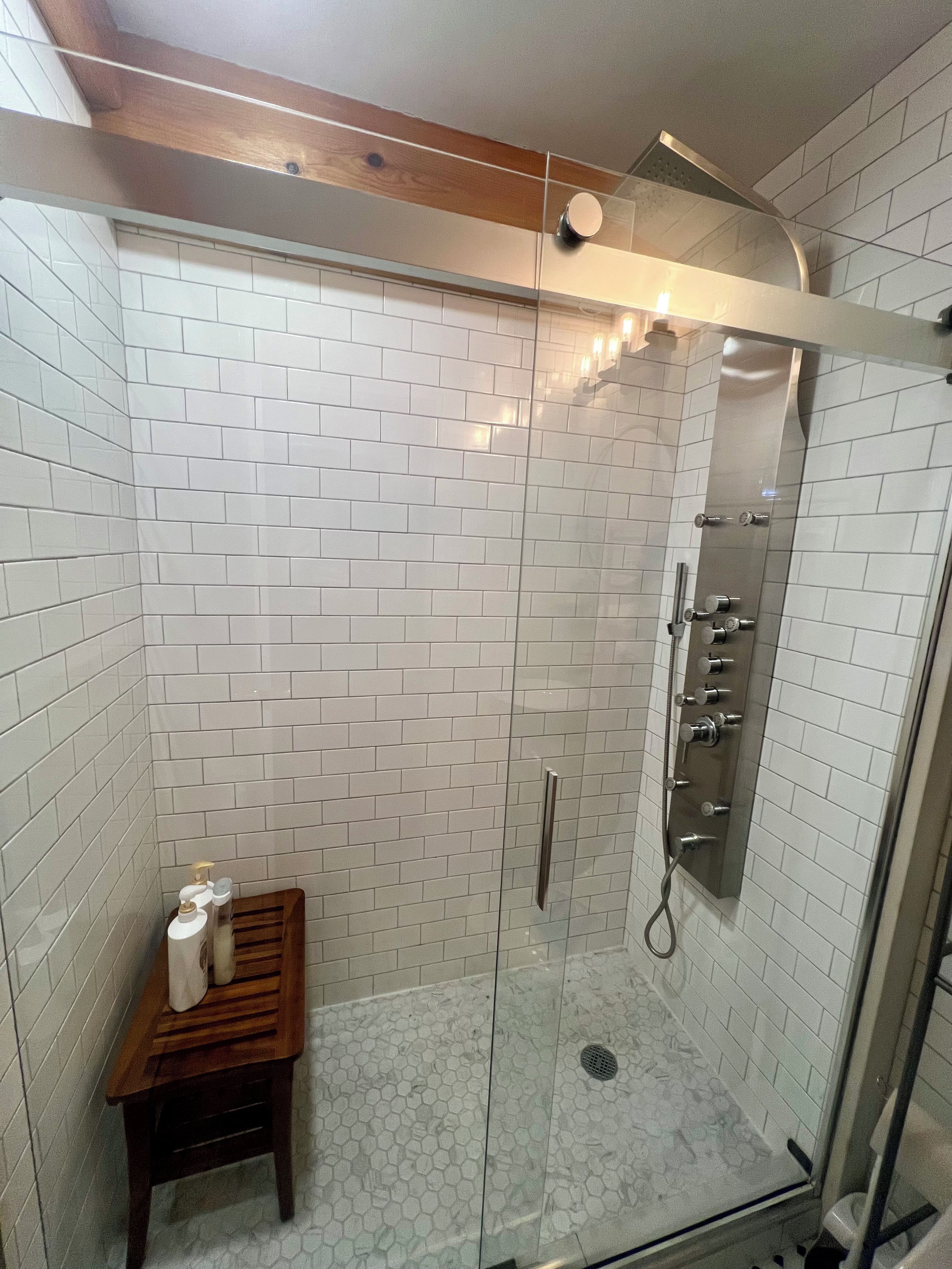 Ducha con cerramiento de vidrio, paredes de azulejos blancos y un banco de madera.