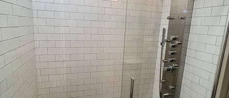 Ducha con cerramiento de vidrio, paredes de azulejos blancos y un banco de madera.