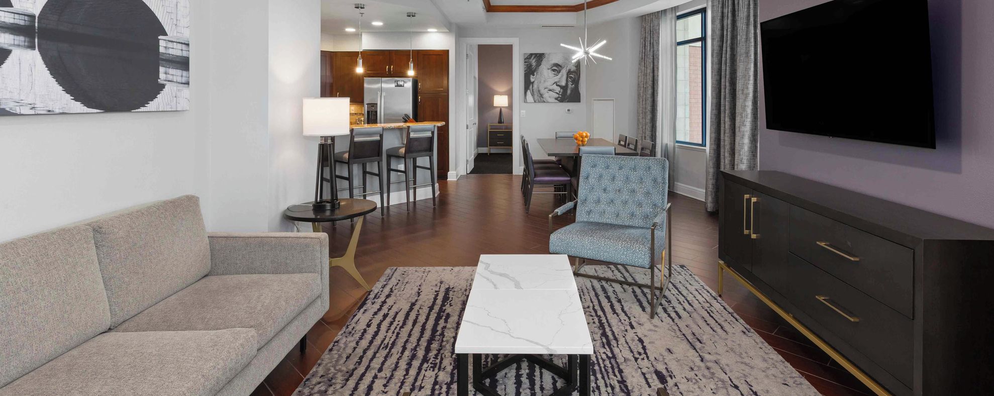 A modern living room with a sofa, a coffee table, and a TV. There is a dining area with chairs and a table, and a kitchen in the background.