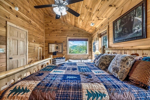 A wooden cabin bedroom with a large bed, a ceiling fan, a window with a view of greenery, and a bear painting on the wall. - Fox Den on Valley View - Hot tub & Fire pit (Pigeon Forge)