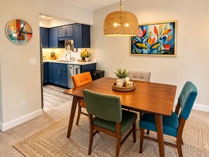 A dining area with a wooden table, four chairs, a woven pendant light, a colorful abstract painting, and a kitchen in the background. - 2-bedroom condo in fabulous Fenton, MI. (Fenton)
