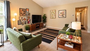 A living room with two green sofas, a flat-screen TV, a wooden cabinet, and a patterned rug. - 2-bedroom condo in fabulous Fenton, MI. (Fenton)