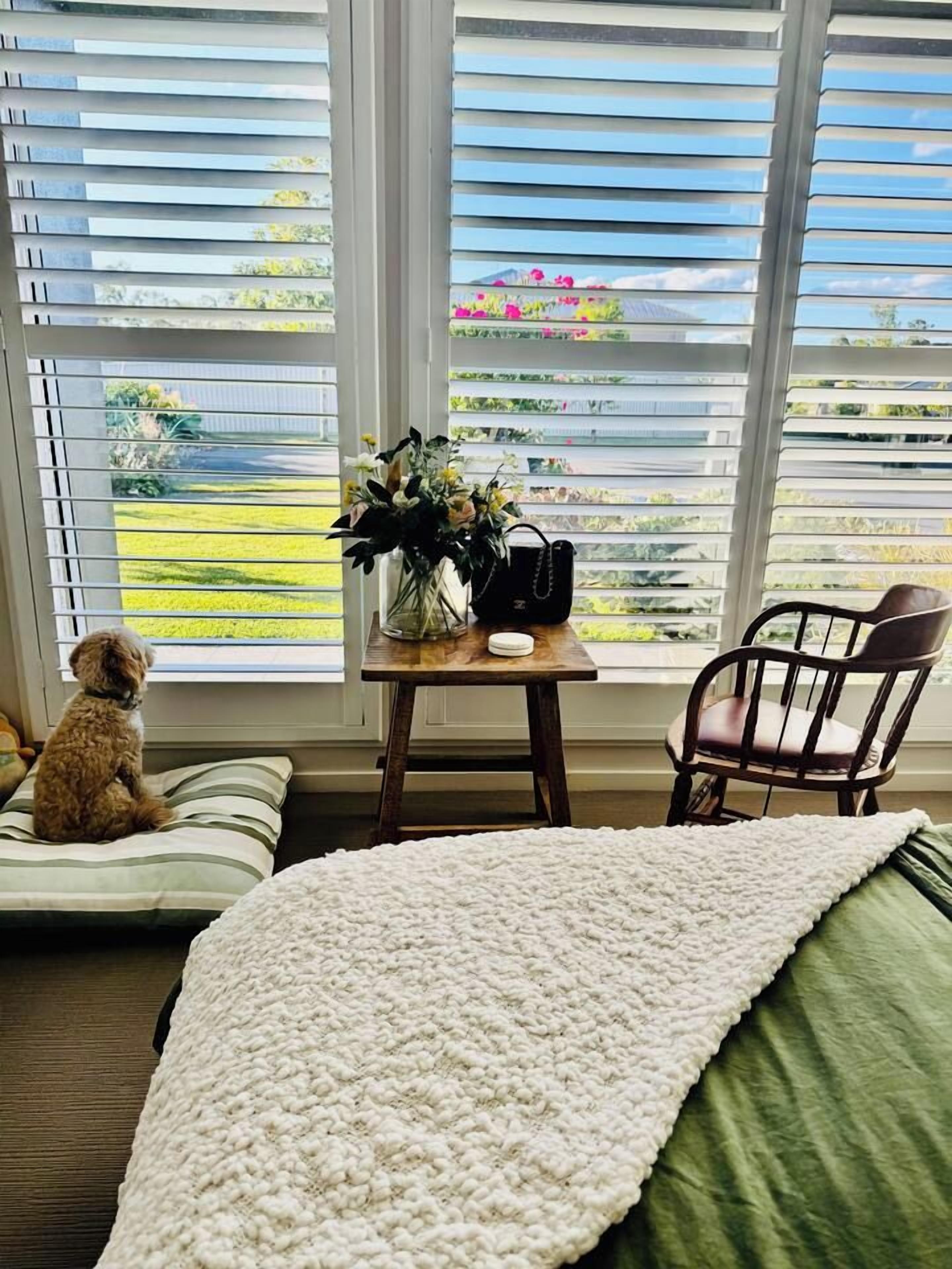 A room with a window, a dog, a chair, a table with a vase, and a bed with a green pillow.