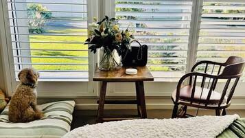 A room with a window, a dog, a chair, a table with a vase, and a bed with a green pillow.