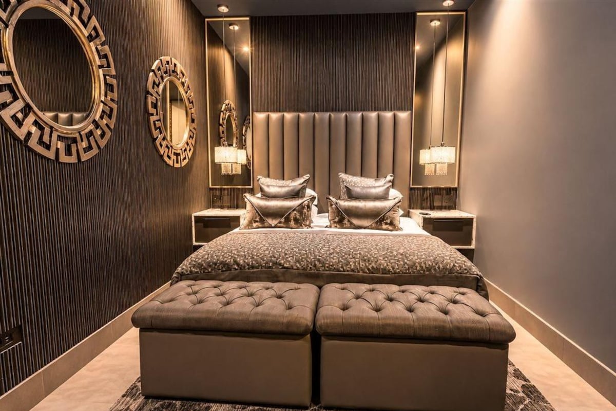 A bedroom with a large bed, two armchairs, a headboard, and wall-mounted lamps.