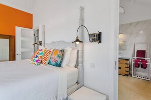 Ein Schlafzimmer mit einem Bett, bunten Kissen, einem Nachttisch, einer Wandlampe und einem im Hintergrund sichtbaren Badezimmer.