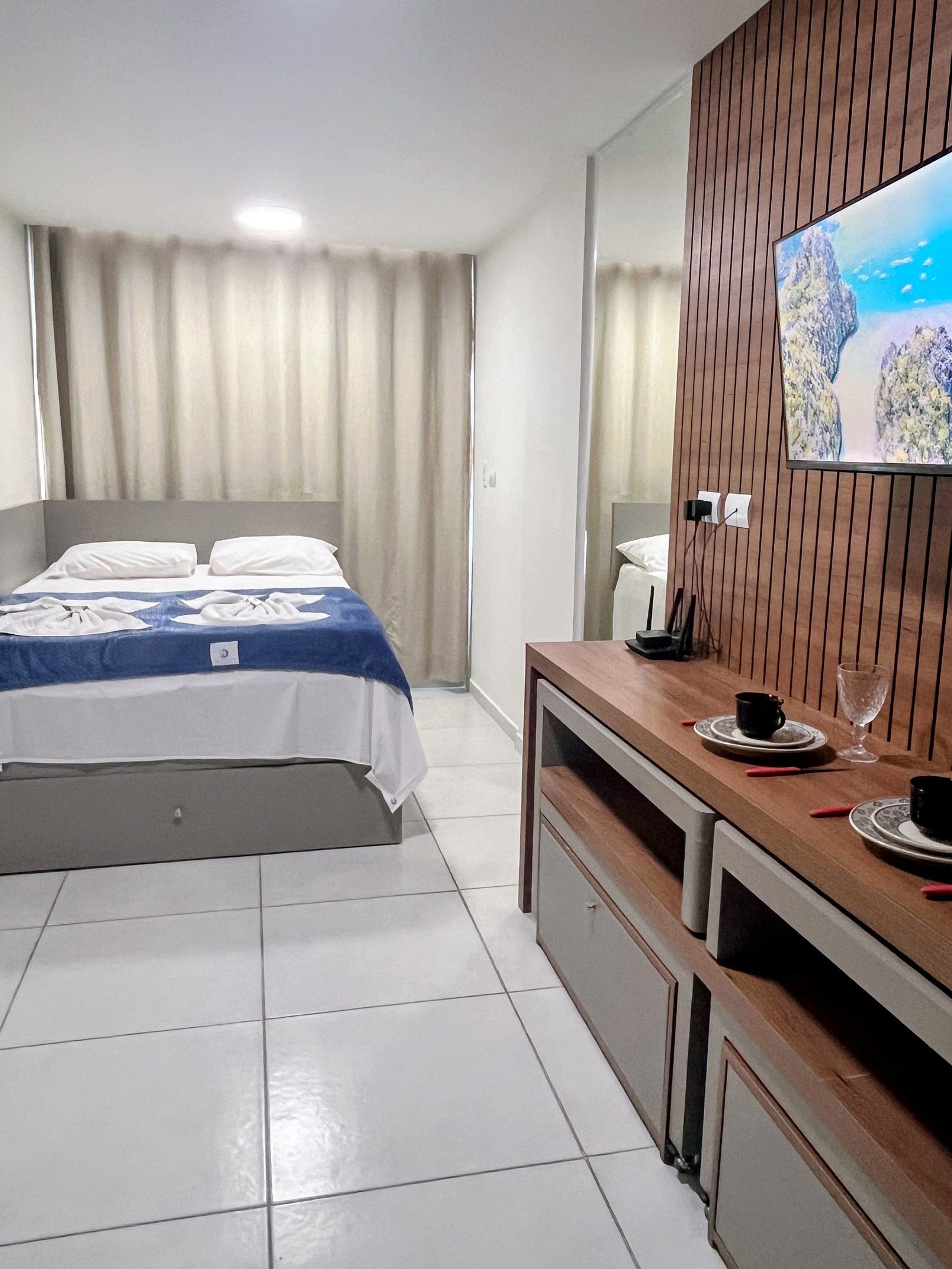 A hotel room with a bed, a TV mounted on a wooden panel, a desk with a cup and saucer, and a window with curtains.
