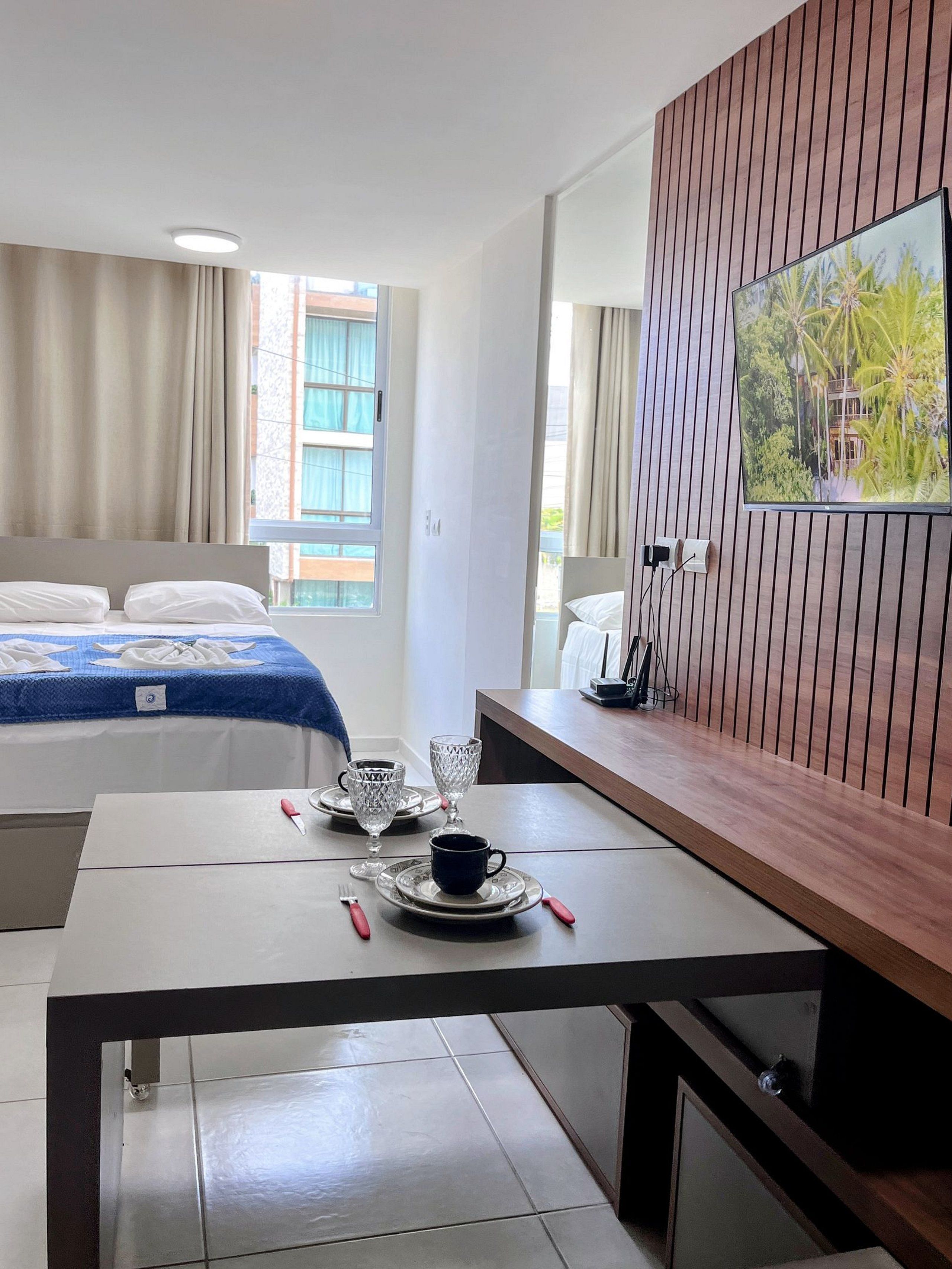 A hotel room with a bed, a dining table with cutlery, a television, and a window with curtains.