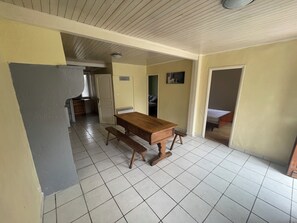 Zona de menjador amb taula i bancs de fusta, cuina al fons i sòl de rajoles.