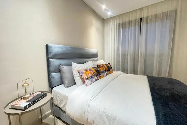 A hotel room with a bed, a side table, and a window with curtains. - Modern 1-Bedroom Apartment in Casablanca | Stylish & Comfortable (Casablanca)