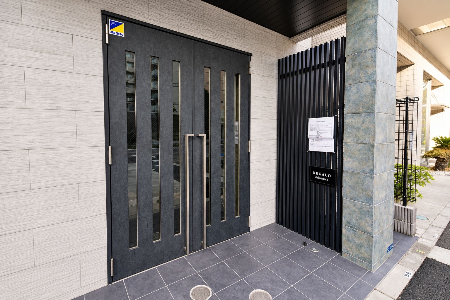 A modern building entrance with a dark door, a vertical metal gate, and a sign indicating 'Regalo shibakura.'