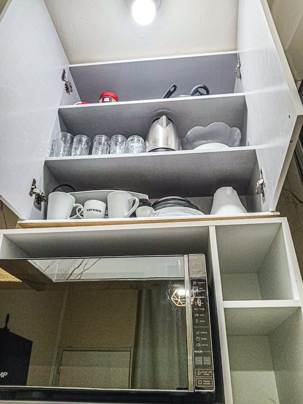 Cuina amb microones, cafetera i una estant plena de vaixella de cuina.