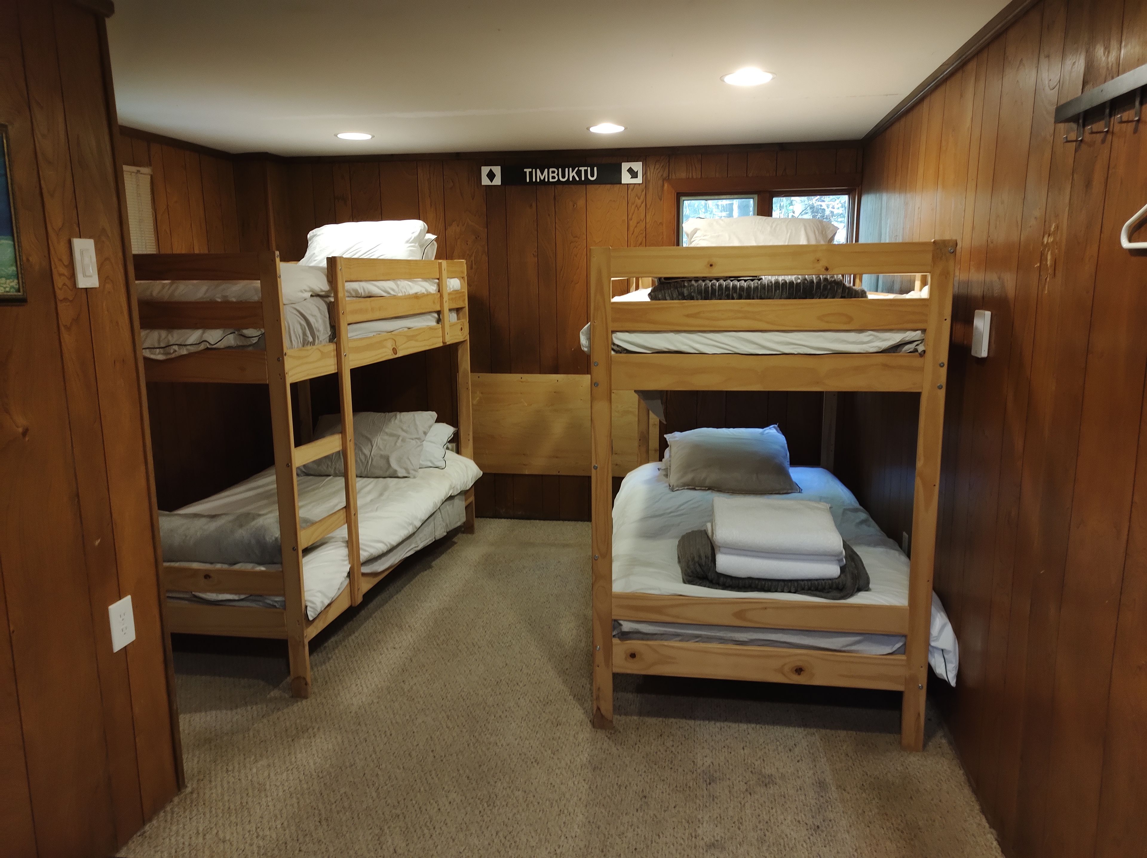 A bunk room with two bunk beds, wooden walls, a sign indicating 'TIMBUKTU,' and a window.