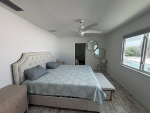 Un dormitori amb llit, taules de nit, ventilador de sostre, finestra i porta que dona a una altra habitació.