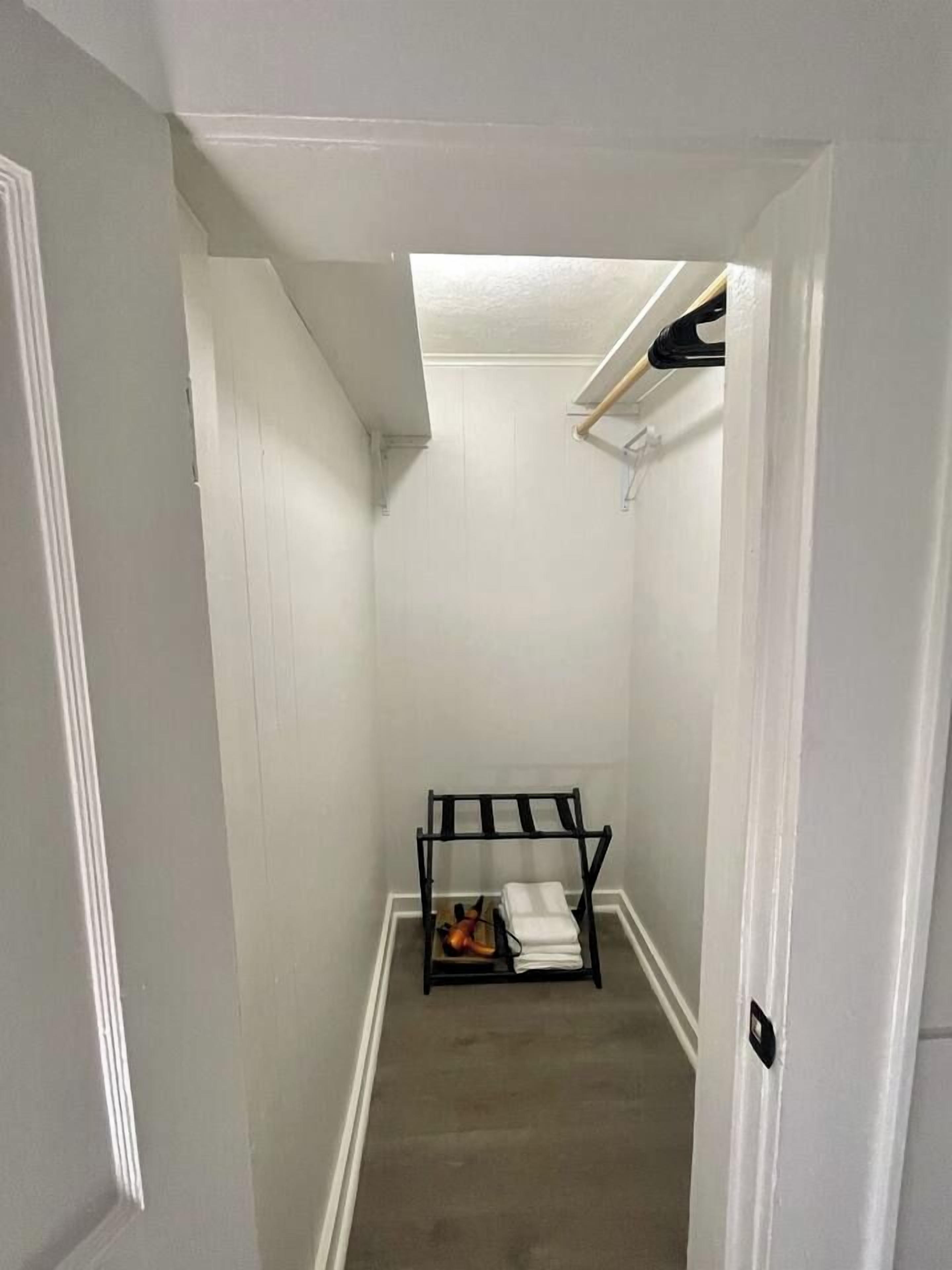 A narrow hallway with white walls and a door on the left. A black rack with folded items is placed against the wall.