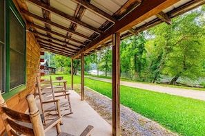 A covered outdoor seating area with wooden benches, a concrete floor, and a view of a green landscape with trees and a path. - Eagle's Nest | Riverfront Cabin Near Bryson City (Swain County)