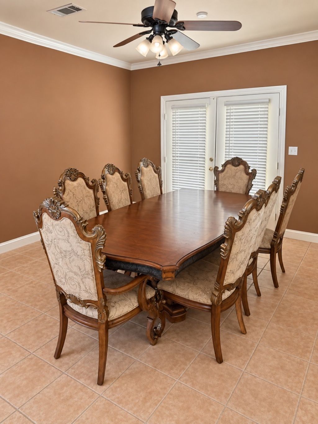 A dining room with a large wooden table surrounded by ornate chairs, a ceiling fan, and a door with white shutters.