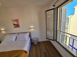 Een hotelkamer met een bed, een klein nachtkastje, een wandlamp en een raam met uitzicht op gebouwen en boten.