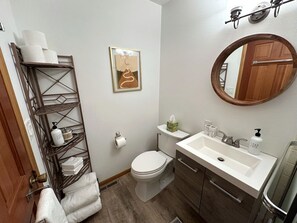 Un baño con inodoro, lavamanos y una unidad de estantería metálica con artículos de aseo.