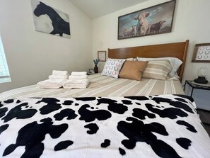 A neatly made bed with a striped bedspread and a cow-patterned blanket, a wooden headboard, and a painting of cows on the wall. - Tobin Hill Casita w/ Private Hot Tub near Pearl (San Antonio)