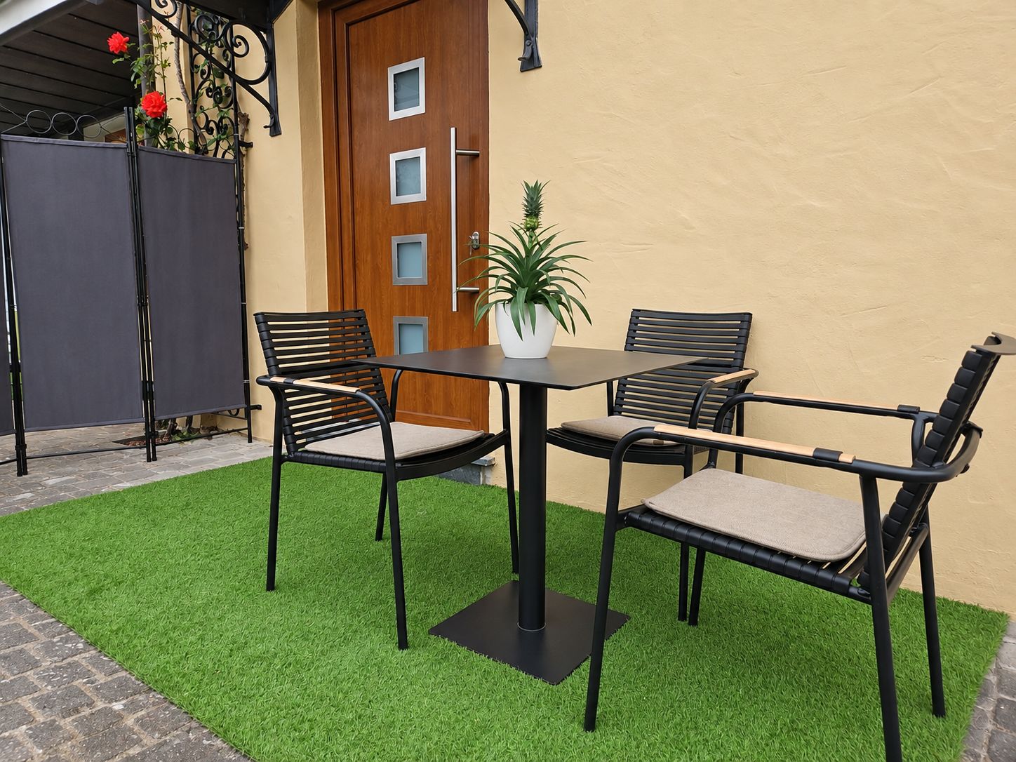 Une terrasse avec une table et quatre chaises, un pot de fleurs et une porte avec une fenêtre.