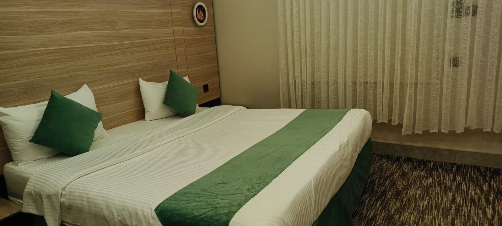 A hotel room with a wooden headboard, a bed with green and white bedding, and a window with sheer curtains.