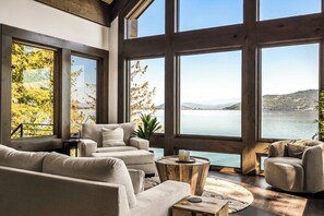 A spacious living room with a large window overlooking a lake, furnished with a sofa and armchairs. - Premier Lakefront Timber Frame Lodge | 2 Kitchens, Bunk Room & Dock | Guest House (Sagle)