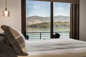 A bedroom with a large window overlooking a lake and hills. - Premier Lakefront Timber Frame Lodge | 2 Kitchens, Bunk Room & Dock | Guest House (Sagle)