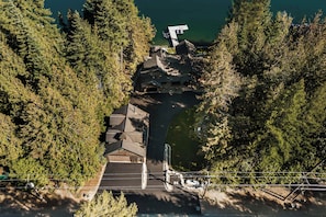 An aerial view of a lodge with a dock extending into a lake, surrounded by dense forest. - Premier Lakefront Timber Frame Lodge | 2 Kitchens, Bunk Room & Dock | Guest House (Sagle)