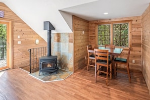 A wooden interior with a fireplace, a dining table with chairs, and a window with a view of greenery. - Premier Lakefront Timber Frame Lodge | 2 Kitchens, Bunk Room & Dock | Guest House (Sagle)