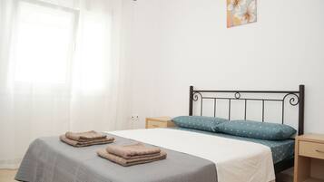 A bedroom with a bed, two bedside tables, a nightstand, a window with sheer curtains, and a wall-mounted artwork.