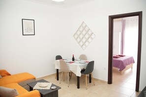 Un comedor con una mesa lista para dos personas, una obra de arte enmarcada en la pared, y una cama visible a través de una puerta abierta.