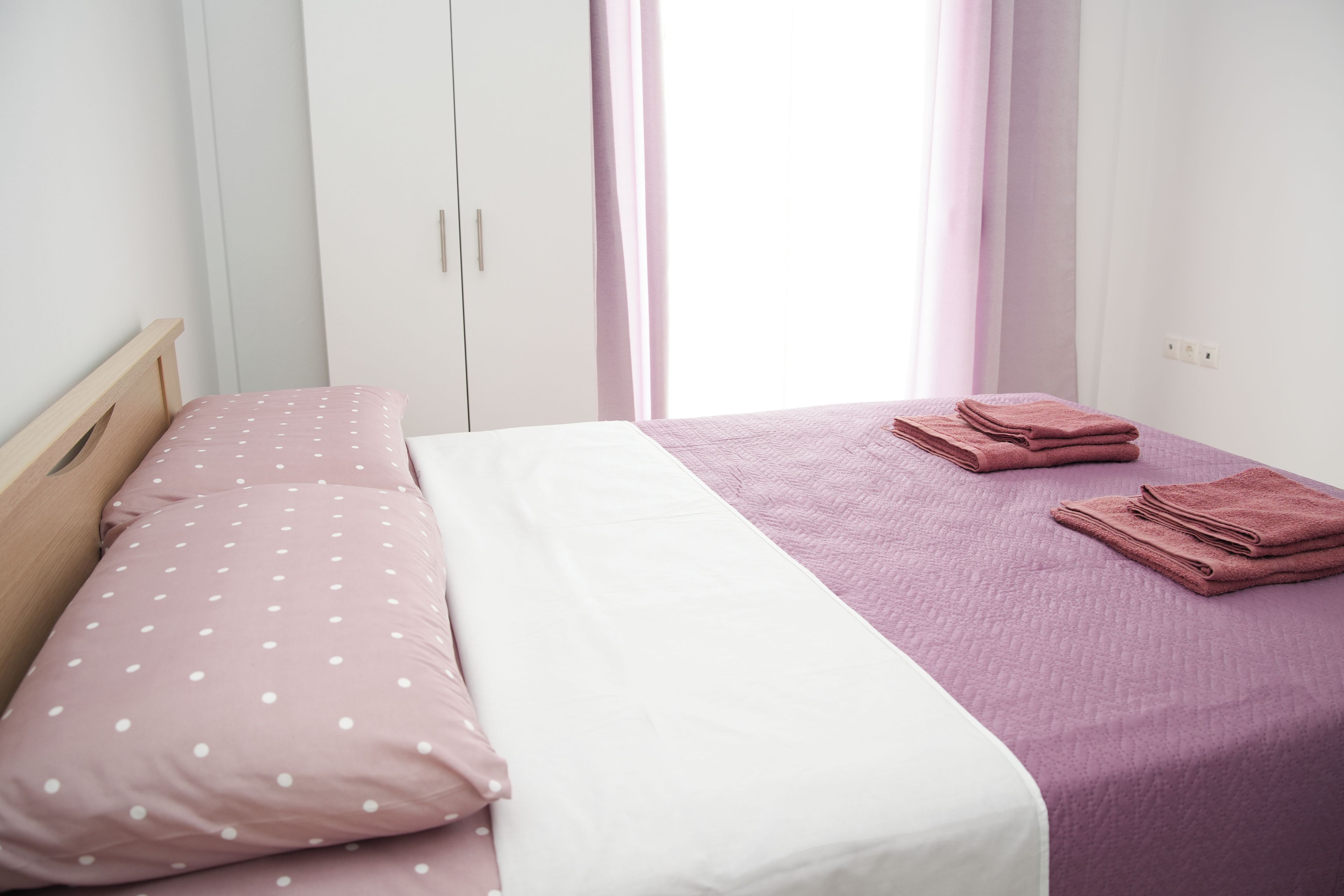 Una cama individual con una colcha blanca y rosa, dos almohadas y una toalla doblada al lado.